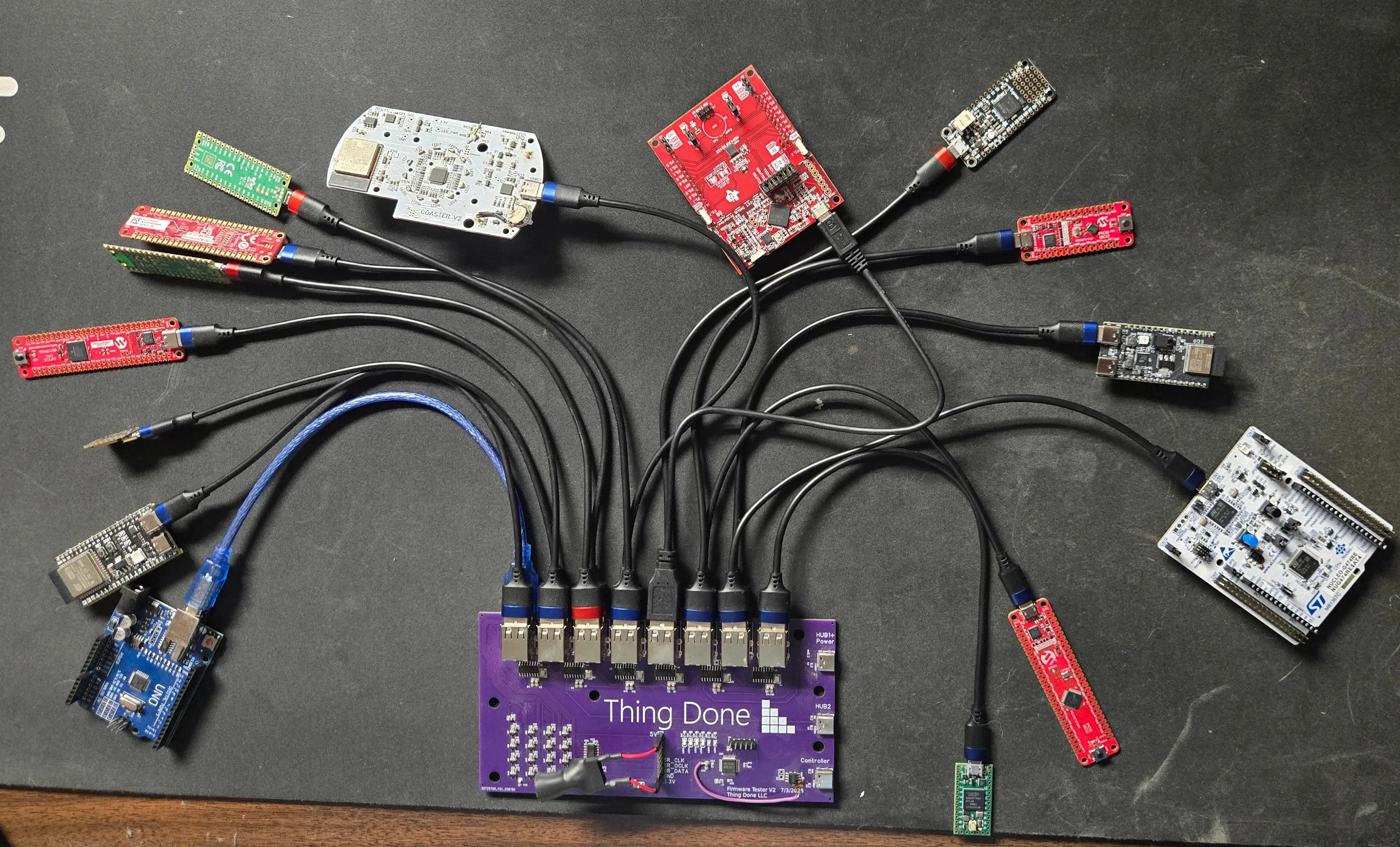 More than a dozen microcontroller dev boards fanned out loose across a dark workbench, every one cabled into a purple carrier PCB with a row of USB ports